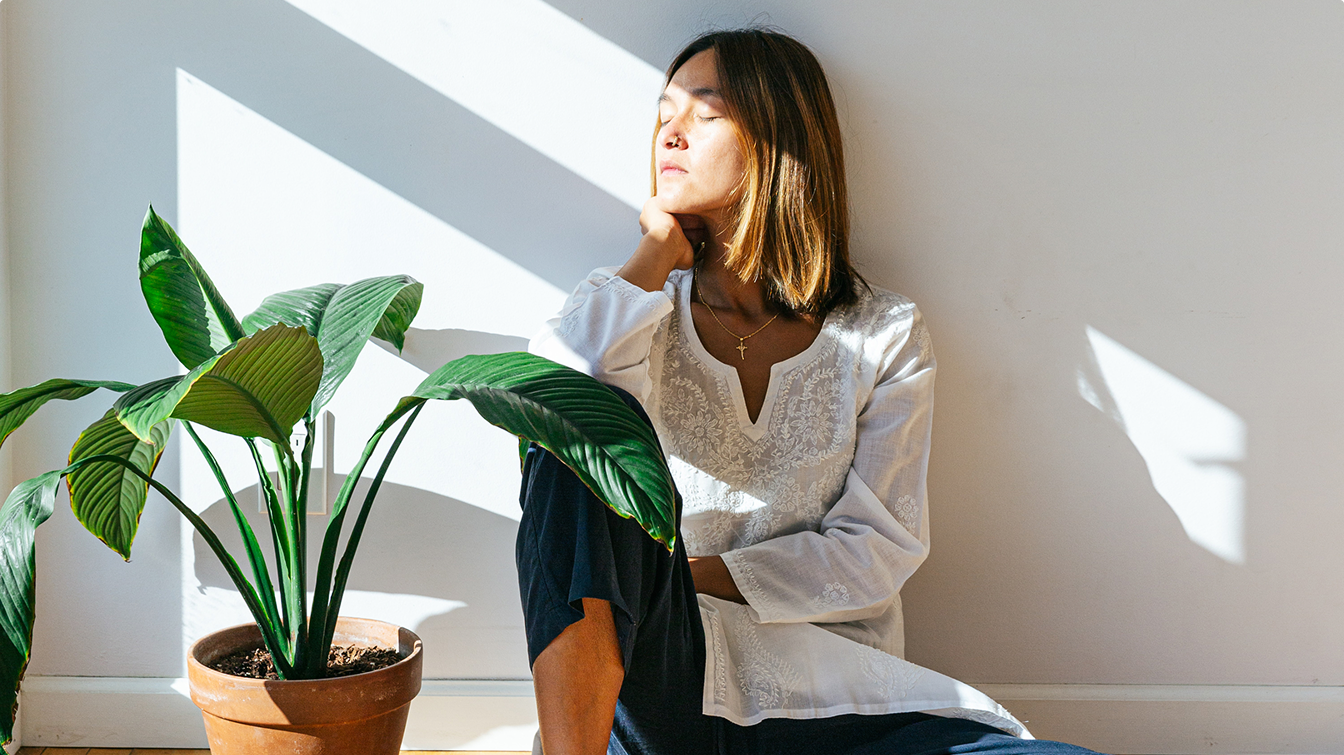 Es ist eine Frau zu sehen, die in ihrem Wohnzimmer auf dem Boden sitzt und aus dem Fenster der Sonne entgegen schaut.
